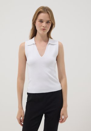 Joven mujer con blusa blanca sin mangas con cuello y escote en V, y pantalones negros, de pie sobre un fondo claro y liso.