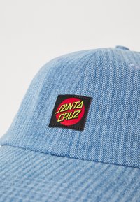 Căciulă din denim albastru, cu o aplicație pătrată neagră și logo-ul roșu și verde "Santa Cruz". Design structurat, cu material texturat și detalii cusute.