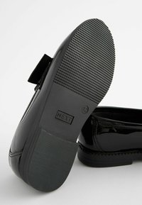 Zwarte gepatineerde leren platte schoenen met een gestructureerde rubberen zool, voorzien van een strikaccent aan de voorkant. Merklabel 'NEXT' zichtbaar op de zool.