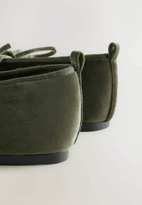 Oliven grønne velours slip-on sko med rundede bageste, struktureret overflade og stofbetrukne løkker ved hælen, hvilende på sorte gummisåler.