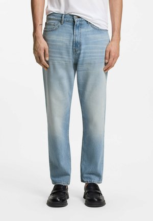 Mężczyzna ubrany w jasnoniebieskie, wyblakłe dżinsy, czarne mokasyny i białą koszulkę, stojący z luźno opuszczonymi rękami na białym tle.