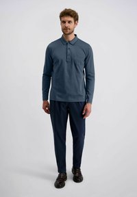 Mann mit kurzen Locken, der ein blaues langärmliges Polohemd, dunkle marineblaue Hose und braune Anzugschuhe trägt, steht vor einem schlichten hellen Hintergrund.