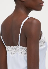 Vaaleansininen satiininen hihaton paita, jossa on pitsireunus kaula-aukossa, ohut säädettävä olkaimet ja kultaiset yksityiskohdat. Sileä pinta, herkkä muotoilu.