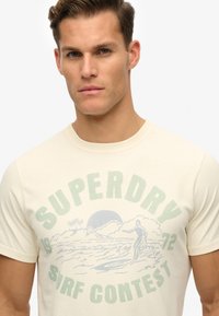 Camiseta de algodón crema con gráfico "SUPERDRY" en verde claro, con escena de surf y palmeras, cuello redondo y mangas cortas.