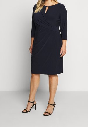 Kobieta ubrana w granatową sukienkę o długości do kolan, z rękawami 3/4 i dekoltem w kształcie łezki, w połączeniu z czarnymi sandałami na wysokim obcasie z paskami.