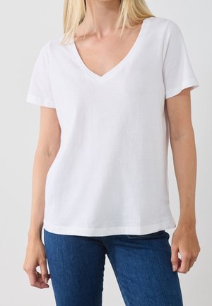 Person iført en ensfarvet hvid kortærmet V-hals T-shirt og blå jeans, vist fra nakke til hofter mod en neutral baggrund.