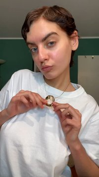 Goudkleurige hangersketting met een glad, ovaal ontwerp en een centrale translucente steen, gedragen aan een dunne ketting, tegen een wit overhemd.