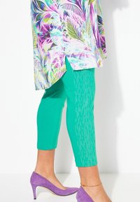 Färgglad blommig tunika med gröna capribyxor som har vertikala linjeaccentuer, kombinerat med lila högklackade skor. Ett lätt och luftigt material används.