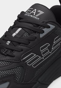Μαύρο αθλητικό παπούτσι με παχιές κορδόνια, πλευρές από δίχτυ και το λογότυπο EA7 Emporio Armani στη γλώσσα και στην πλευρική επιφάνεια με λευκές και γκρι λεπτομέρειες.
