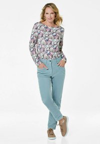 Een blouse met lange mouwen met een bloemenpatroon in tinten roze en blauw, gecombineerd met lichtblauwe broek en bruine sneakers, staand tegen een witte achtergrond.