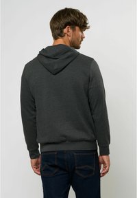 Hanorac gri închis cu glugă ajustabilă, manșete și tiv cu nervuri. Țesătura pare moale cu o textură netedă. Purtați cu blugi albaștri cu buzunare la spate.