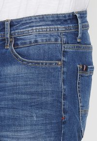 Jeans de mezclilla azul con un diseño clásico, que presentan un estilo de cinco bolsillos, un ligero desvanecimiento y un acento rojo en la costura del bolsillo trasero.