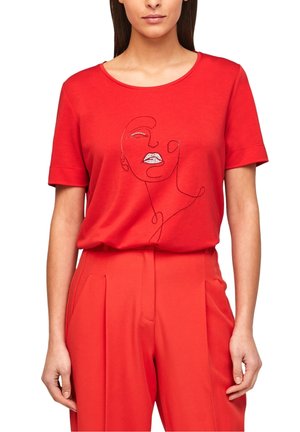 Femme portant un t-shirt rouge à manches courtes avec un dessin de visage en lignes noires et un pantalon rouge assorti sur fond blanc.