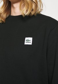 Žena nosící černou mikinu Lacoste se zeleným logem krokodýla na hrudi a s viditelnými blond vlasy na bílém pozadí.