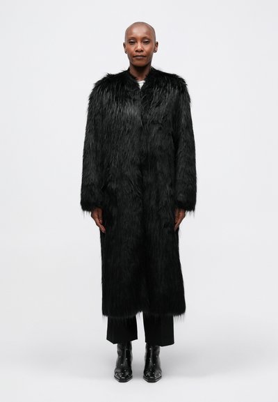 Manteau en fausse fourrure noir avec des manches longues, doté d'une texture en peluche et d'une coupe droite, porté sur un pantalon noir et des bottines vernies.