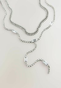 Stříbrný náhrdelník se třemi prameny, který obsahuje kulaté a obdélníkové čiré rhinestony uspořádané v vlnitém vzoru. Hladký metalický povrch.