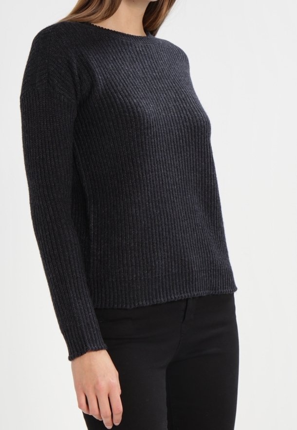 Femme portant un pull en maille côtelée noir ajusté à manches longues et un pantalon noir sur un fond uni.