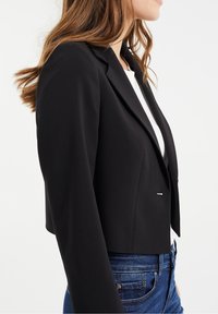 Black cropped blazer with a structured design, notched lapels, and a single silver accent button. Worn over a white top and blue jeans.