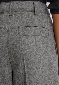 Close-up van grijze gestructureerde broek met een achterzak, gedragen met een zwarte geribbelde mouw zichtbaar aan de rechterkant.