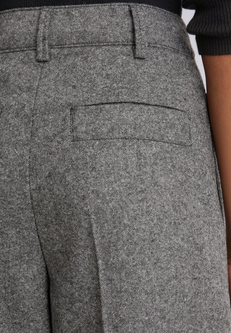 Close-up van grijze gestructureerde broek met een achterzak, gedragen met een zwarte geribbelde mouw zichtbaar aan de rechterkant.