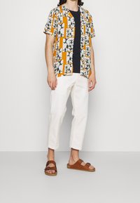 Hemd mit kurzen Ärmeln in einem weißen, schwarzen und orangefarbenen geometrischen Muster; kombiniert mit weißen Hosen und braunen Sandalen.