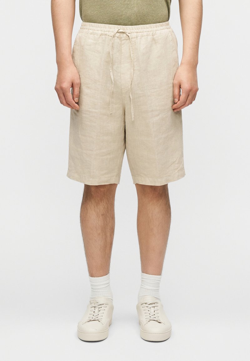 Μπεζ λινά σορτς με ελαστική μέση και κορδόνι. Έχο υν χαλαρή εφαρμογή και φτάνουν λίγο πάνω από τα γόνατα. Συνδυάζονται με λευκά αθλητικά παπούτσια.