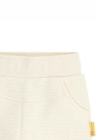 Cremefarbene Kinderhose aus strukturiertem Stoff, mit elastischem Bund, Seitentasche und einem kleinen gelben Etikett auf der rechten Seite.