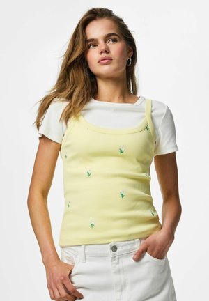 Jeune femme aux longs cheveux bruns portant un t-shirt blanc côtelé et un débardeur jaune avec une petite broderie de fleur blanche, les mains dans les poches d’un pantalon blanc.
