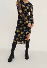 Schwarzes Blumenmusterkleid mit langen transparenten Ärmeln und seitlichem Schlitz. Mit bunten gelben und blauen Blumenmustern. Kombiniert mit kniehohen schwarzen Stiefeln.