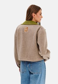 Brązowa kurtka polarowa z zielonym ściągaczem, z kwadratową naszywką z grafiką tematyczną o jedzeniu na plecach. Niebieskie dżinsy dopełniają outfit.