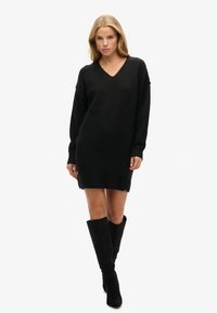 Vestido de punto negro con escote en V, mangas largas y corte relajado, dobladillo acanalado, combinado con botas negras hasta la rodilla.