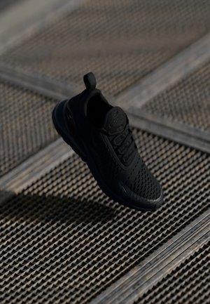 Black athletic sneaker with a perforated upper, textured sole, and a pull tab at the heel, displayed against a patterned surface.