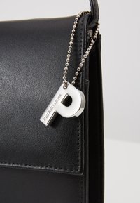 Zwarte leren handtas met gladde textuur, voorzien van een zilverkleurig metalen label in de vorm van de letter 'P', bevestigd met een kraalscharnier.