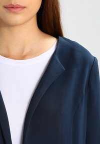Marineblauwe blazer met een gladde textuur en scherpe revers, gedragen over een witte ronde hals t-shirt, met een close-up van de halslijn.