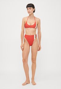 Sarkana bikini ar trīsstūra augšu, kurai ir plānas siksniņas un augsto jostas vietu, izceļot daudzkrāsainas svītras sānos.