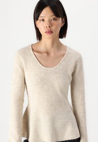 Pull en ribbed beige clair avec un large col rond, des manches longues et un ourlet légèrement évasé. Le matériau semble doux et texturé.
