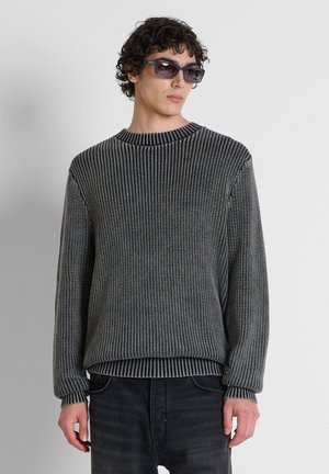 Giovane uomo con capelli ricci che indossa occhiali da sole scuri, un maglione a maglia bianco e nero e pantaloni neri su uno sfondo chiaro e uniforme.