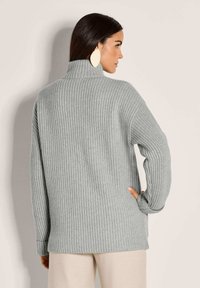 Frau, die einen hellgrauen, gerippten Rollkragenpullover und beige Hosen trägt, seitlich steht und mit einer Hand auf der Hüfte.