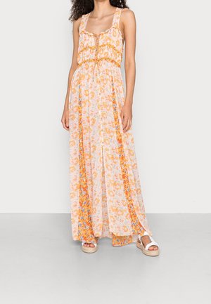 Femme portant une robe longue sans manches à fleurs orange et blanche avec une fente à l'avant, associée à des sandales à plateforme blanches.