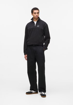 Man standing with one hand in pocket wearing black zip-up fleece, black pants, and black-and-white sneakers on white background.