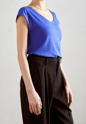 Personne portant un haut bleu manches courtes en V rentré dans un pantalon plissé noir taille haute avec une bague en or à la main gauche.