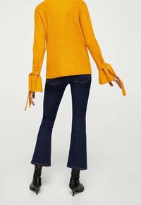 Kobieta w musztardowym swetrze z rozszerzanymi rękawami, związanymi na nadgarstkach, ciemnoniebieskich dzwonach oraz czarnych botkach na obcasie, pokazana od tyłu.