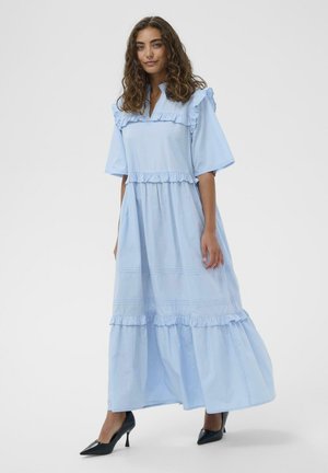 Kihara juustega naine, kes kannab helesinist rööpitud maksikleit ja musti teravate kontsadega kingi, seisab ühevärvilise tausta ees.