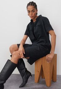 Schwarzes Leinenhemdkleid mit kurzen Ärmeln, Knopfleiste und zwei Brusttaschen. Getragen mit knielangen schwarzen Lederstiefeln, sitzend auf einem Holzstuhl.