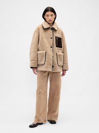 Casaco de pêlo sintético bege com gola, grandes bolsos frontais e um bolso lateral com fecho. Combinado com calças de perna larga castanho claro e sapatilhas pretas.