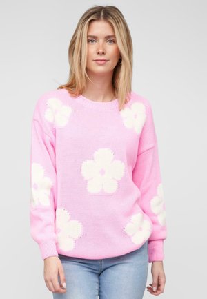 Roze gebreide trui met grote witte bloempatronen, geribbelde manchetten en een ronde halslijn. De stof lijkt zacht en gestructureerd.