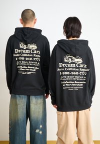 Black hoodies with white text and car graphic on the back; paired with loose denim and beige pants; relaxed fit and casual design.
