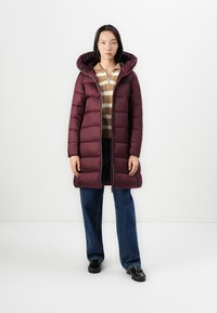 Bordeaux pufferjack met capuchon, voorzien van een ritssluiting aan de voorkant, horizontaal gestikt ontwerp en een lengt tot halverwege de dijen. Gekleed met een gestreept topje en wijde jeans.