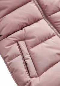 Gilet rembourré en rose clair, avec des sections matelassées, une poche latérale cachée et une texture de tissu lisse et brillante avec une fermeture éclair.