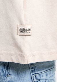 Lichtroze katoenen t-shirt met een wit "Petrol Ind" label gestikt aan de zoom, met blauwe denim jeans op de achtergrond.